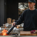 Person grilling outdoors using a Looftlighter with a wooden cutting board and meat on a grill.